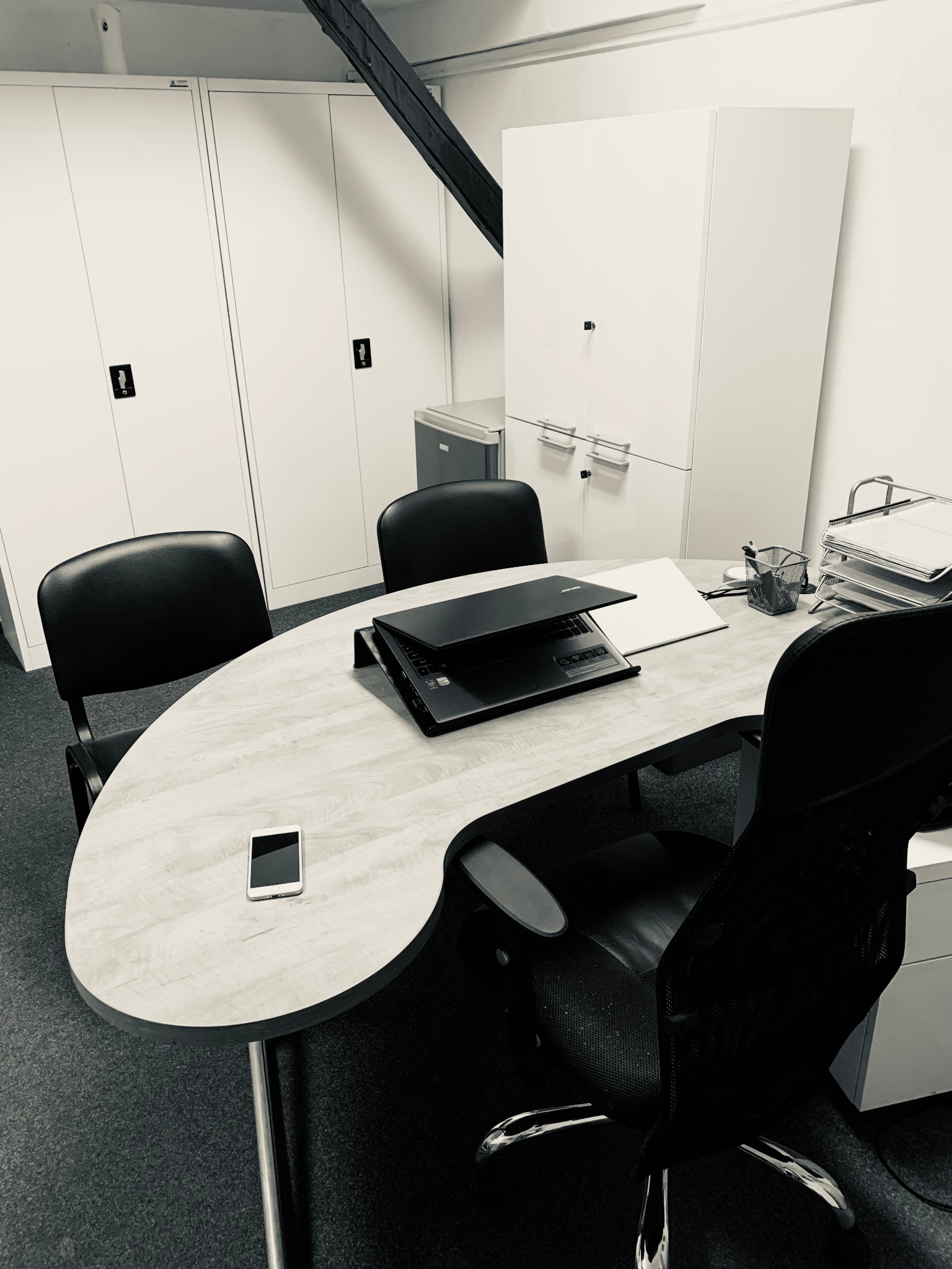 Monochromatyczne zdjęcie biura: owalny stół z laptopem i smartfonem, czarne krzesła, białe szafy w tle. Minimalistyczny wystrój, sugerujący uporządkowane miejsce pracy.