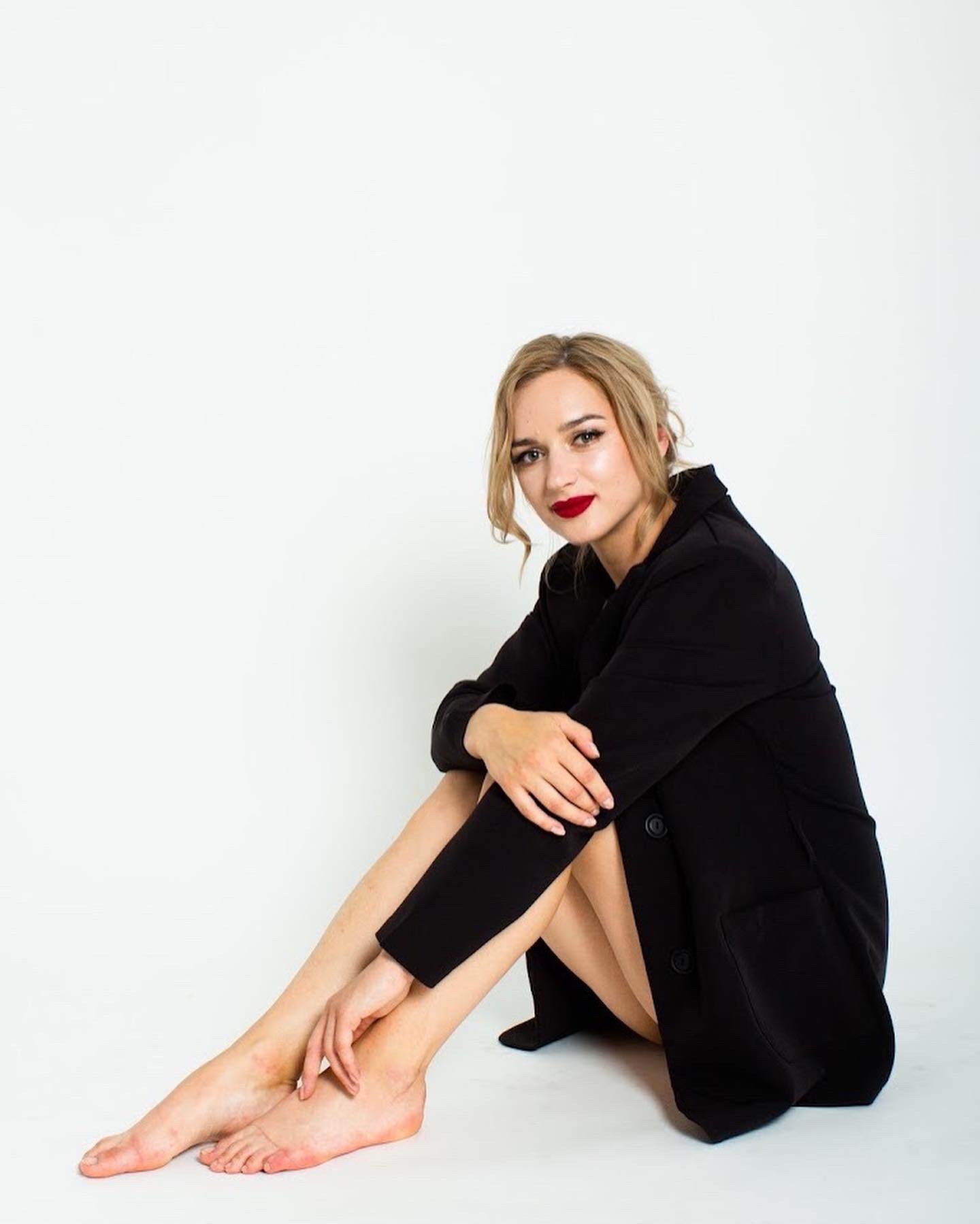 Portret kobiety siedzącej na białym tle, ubrana w czarną marynarkę, z czerwonymi ustami i włosami upiętymi w niedbały kok.