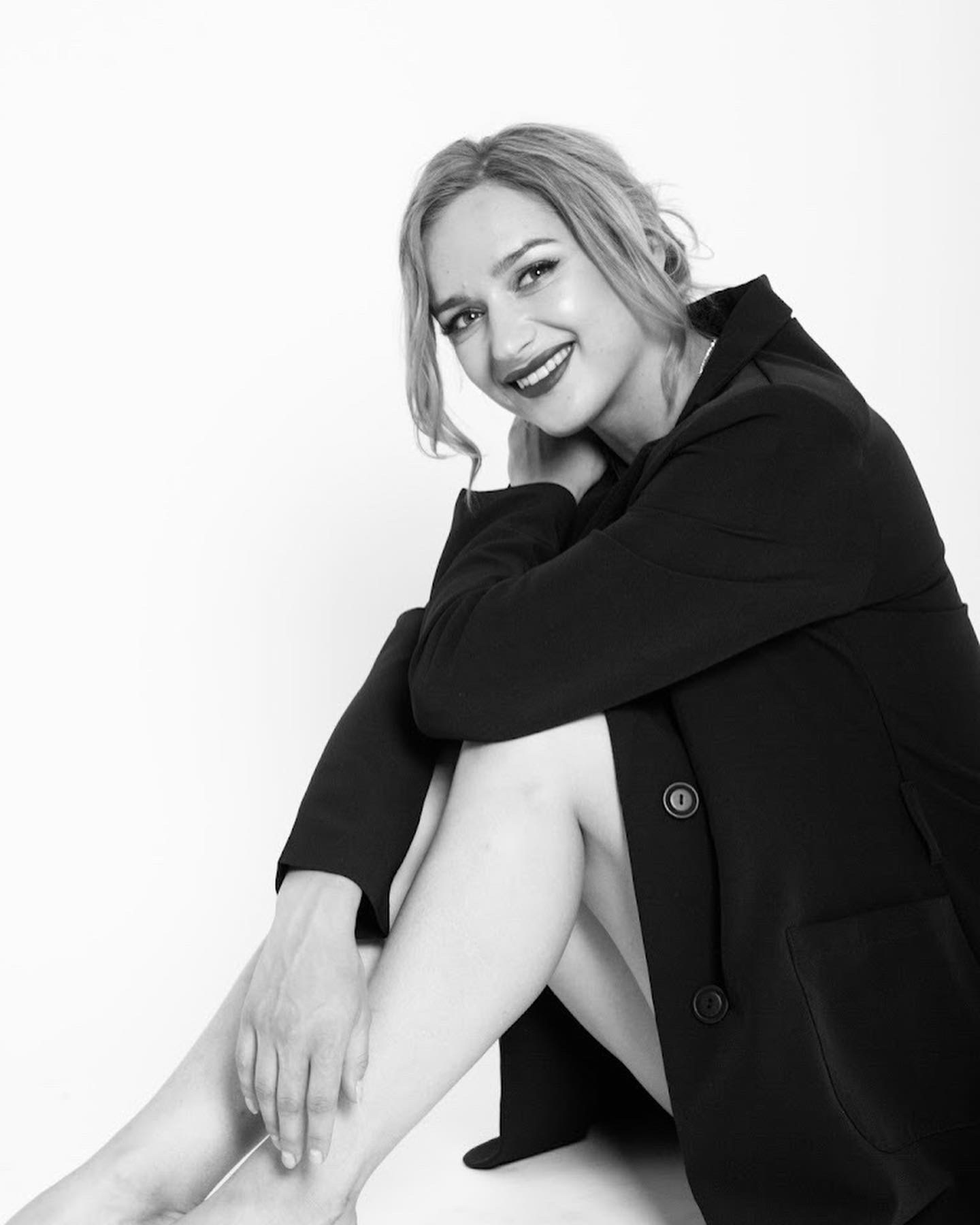 Portret kobiety w czarnej marynarce siedzącej na białym tle, uśmiechniętej, w czarno-białej tonacji.