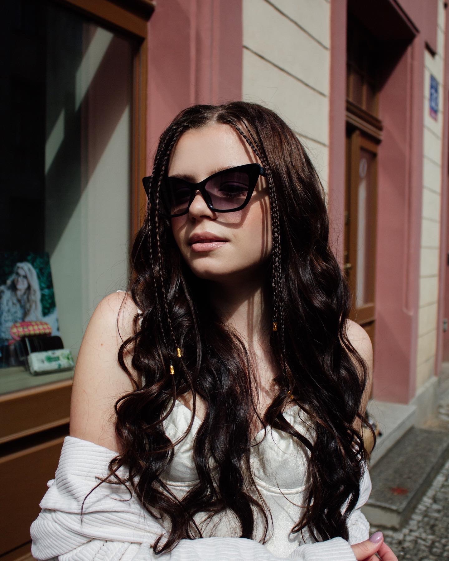 Portret kobiety w okularach przeciwsłonecznych, z długimi, kręconymi włosami z wplecionymi warkoczykami, pozującej na tle budynku z różową fasadą i witryną sklepową.