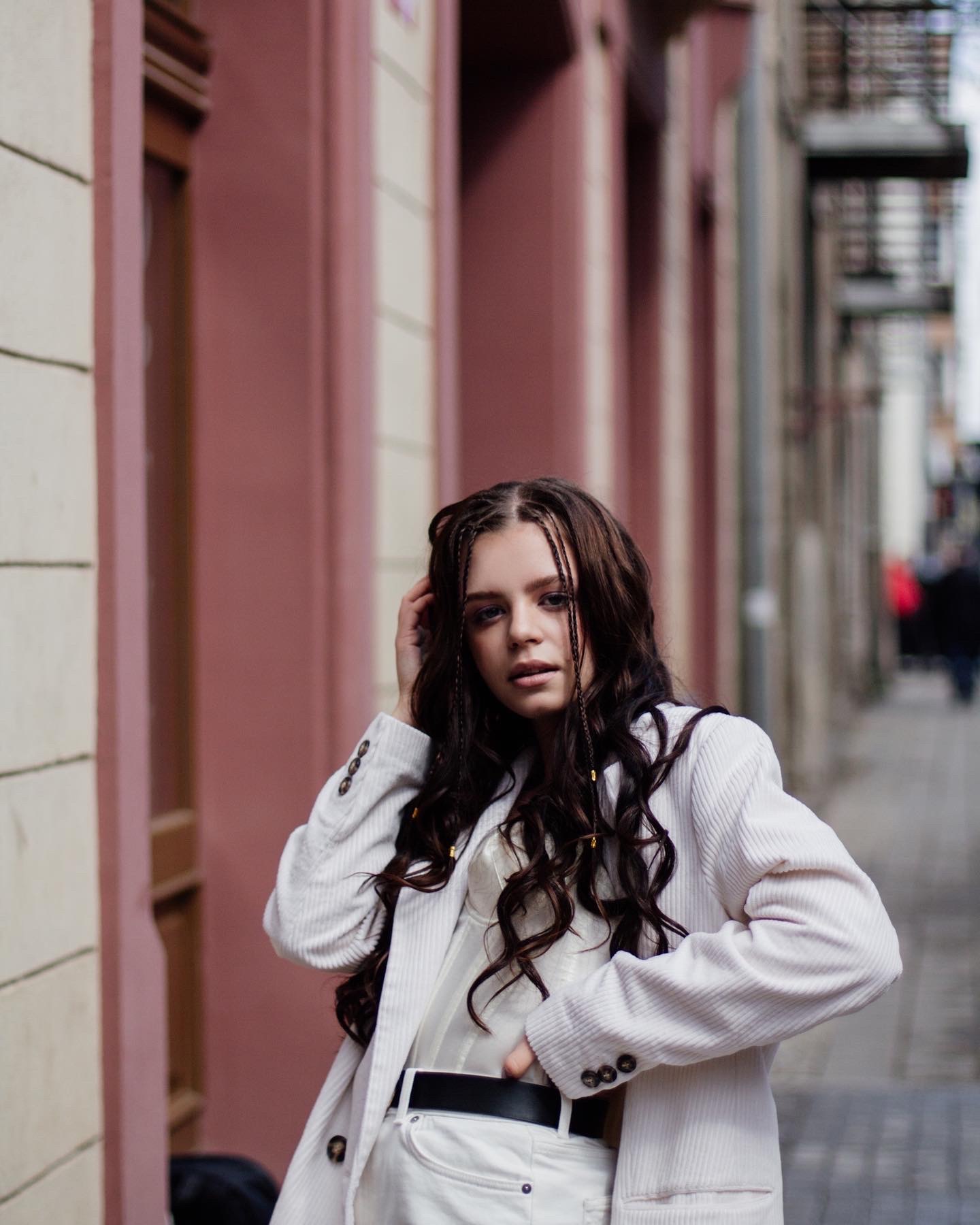 Portret młodej kobiety w białym garniturze sztruksowym na tle miejskiej uliczki z kolorowymi budynkami, z warkoczykami we włosach i delikatnym makijażem.