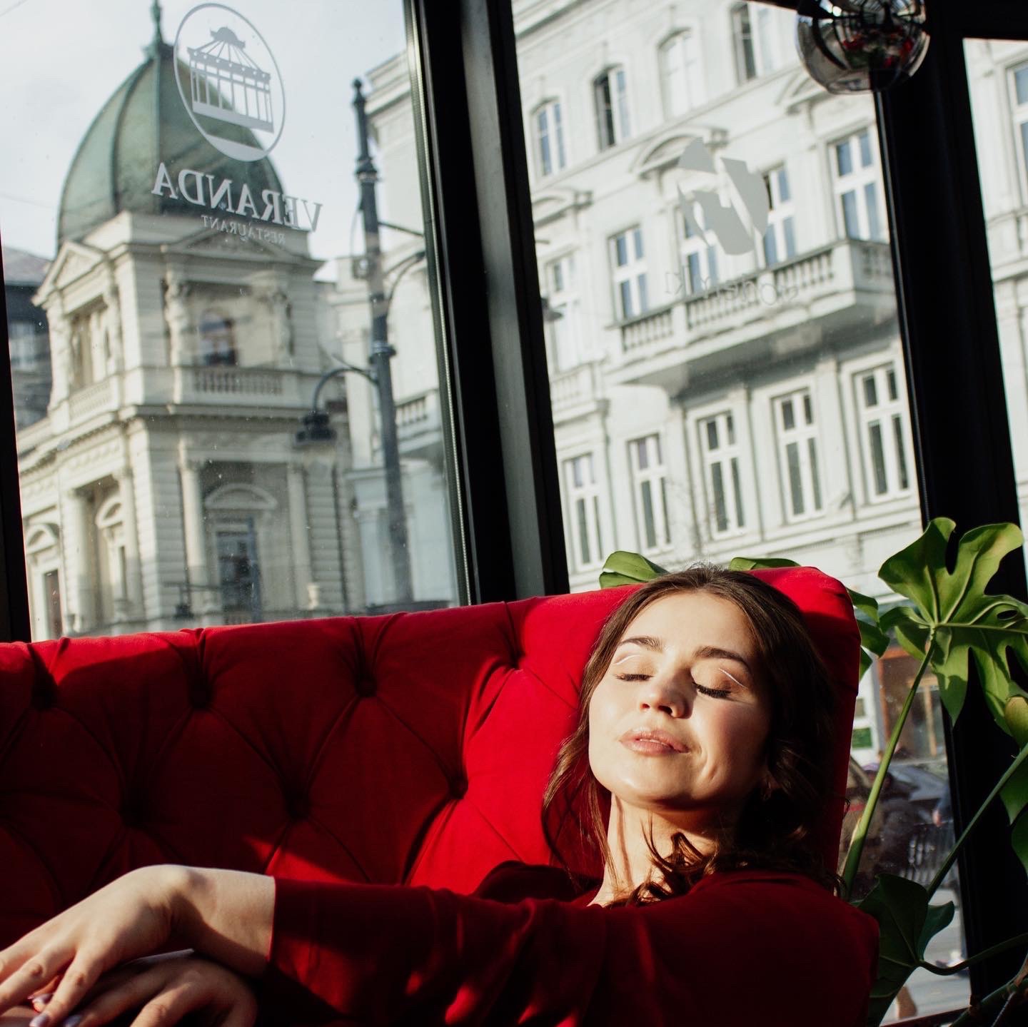 Kobieta z zamkniętymi oczami opiera się na czerwonej, pluszowej sofie w pobliżu okna z widokiem na budynek z kopułą i logo 'Veranda'.