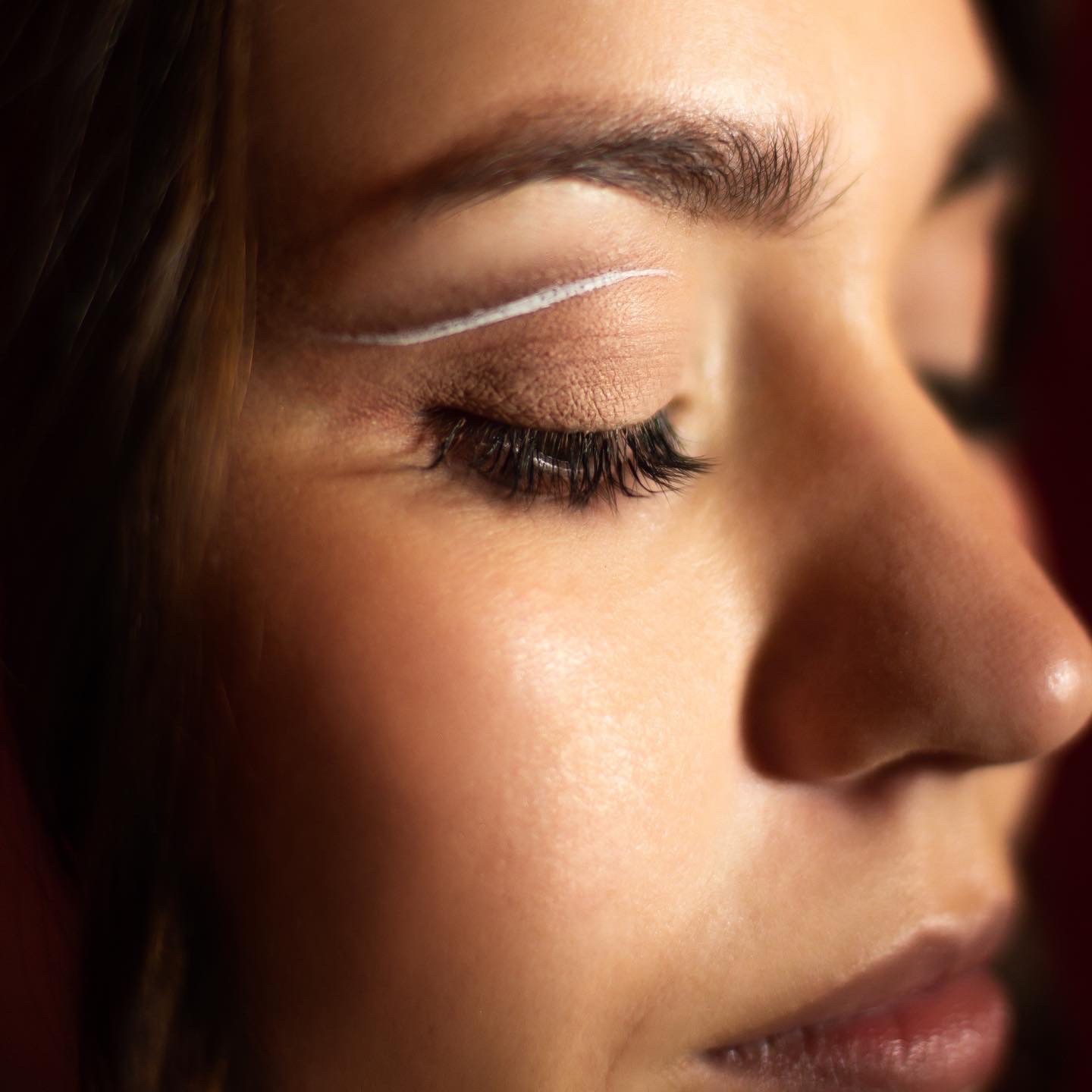 Portret zbliżeniowy oka kobiety z białym eyelinerem w Łodzi, skóra oświetlona ciepłym światłem, widoczne długie rzęsy i delikatny makijaż.