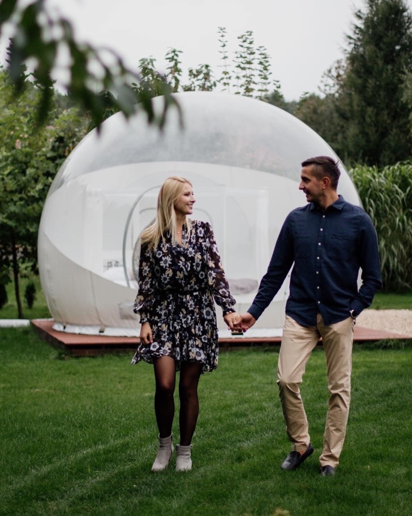 Uśmiechnięta para trzymająca się za ręce spaceruje po trawniku przed przezroczystą kopułą glampingową, kobieta w kwiecistej sukience i botkach, mężczyzna w granatowej koszuli i beżowych spodniach.