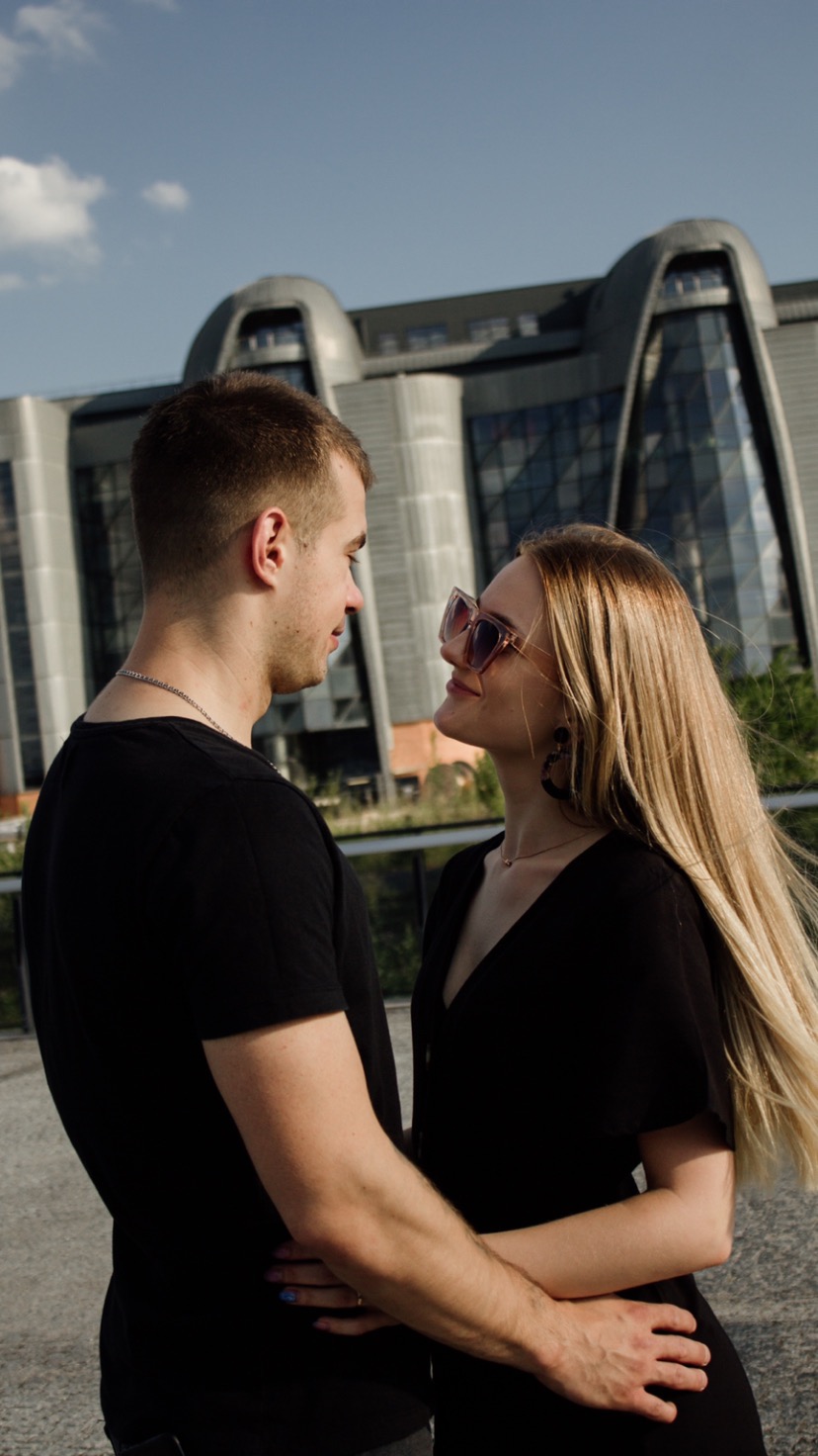 Zakochana para w czarnych ubraniach, ona w okularach, z nowoczesnym budynkiem w tle, wpatrzona w siebie.