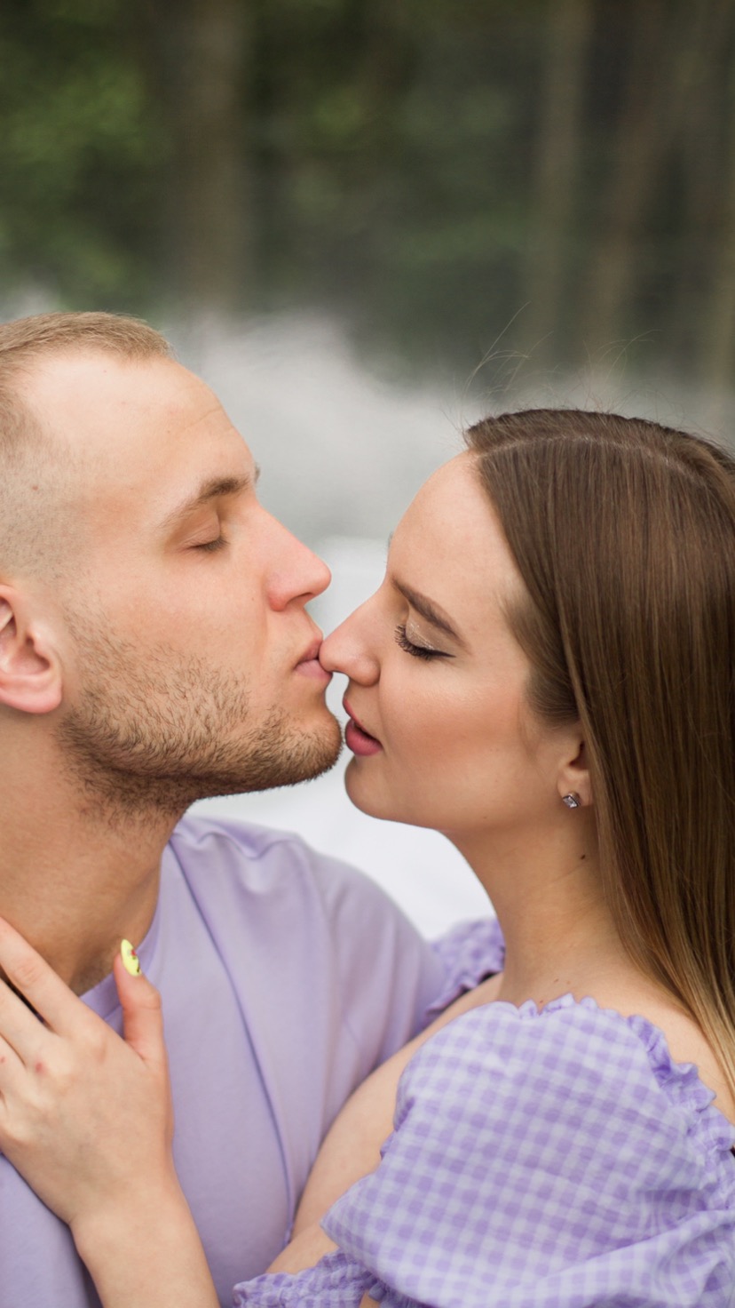 Profil zbliżenie całującej się pary; mężczyzna z zamkniętymi oczami i krótką fryzurą, kobieta z długimi włosami i delikatnym makijażem, oboje ubrani w odcienie fioletu na rozmytym tle.