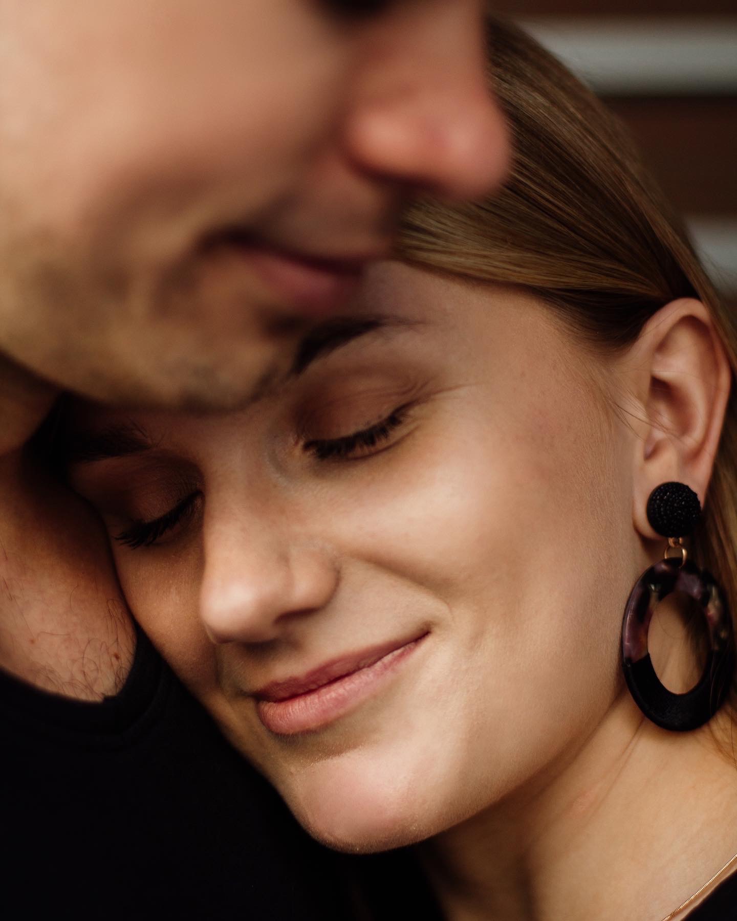 Uśmiechnięta blondynka z zamkniętymi oczami, opierająca głowę o ramię mężczyzny, widoczne kolczyki w kształcie koła z czarną kulką