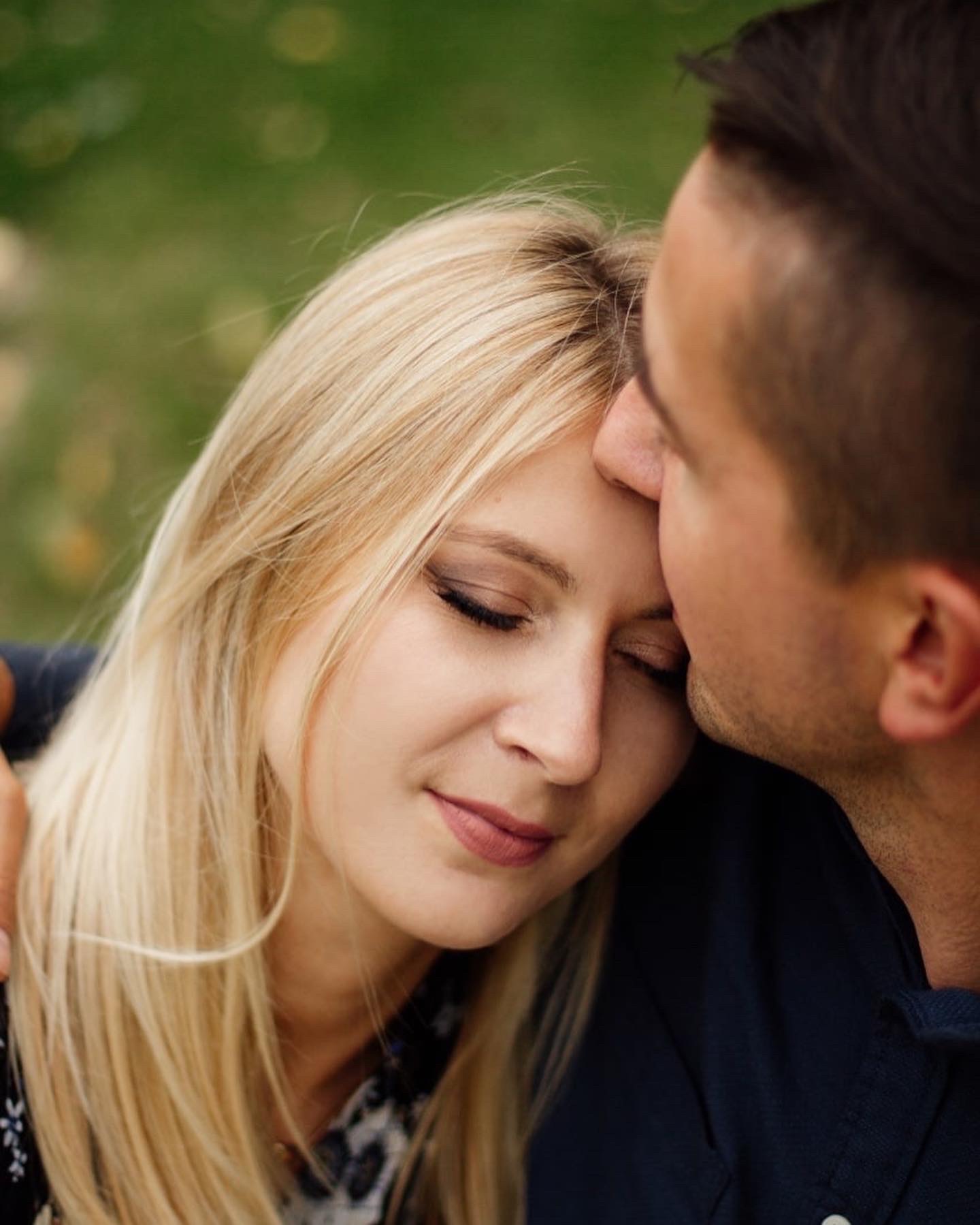 Bliski portret zakochanej pary: mężczyzna całuje kobietę w czoło, ona z zamkniętymi oczami, delikatny uśmiech, blond włosy opadają na ramiona, zielone tło.