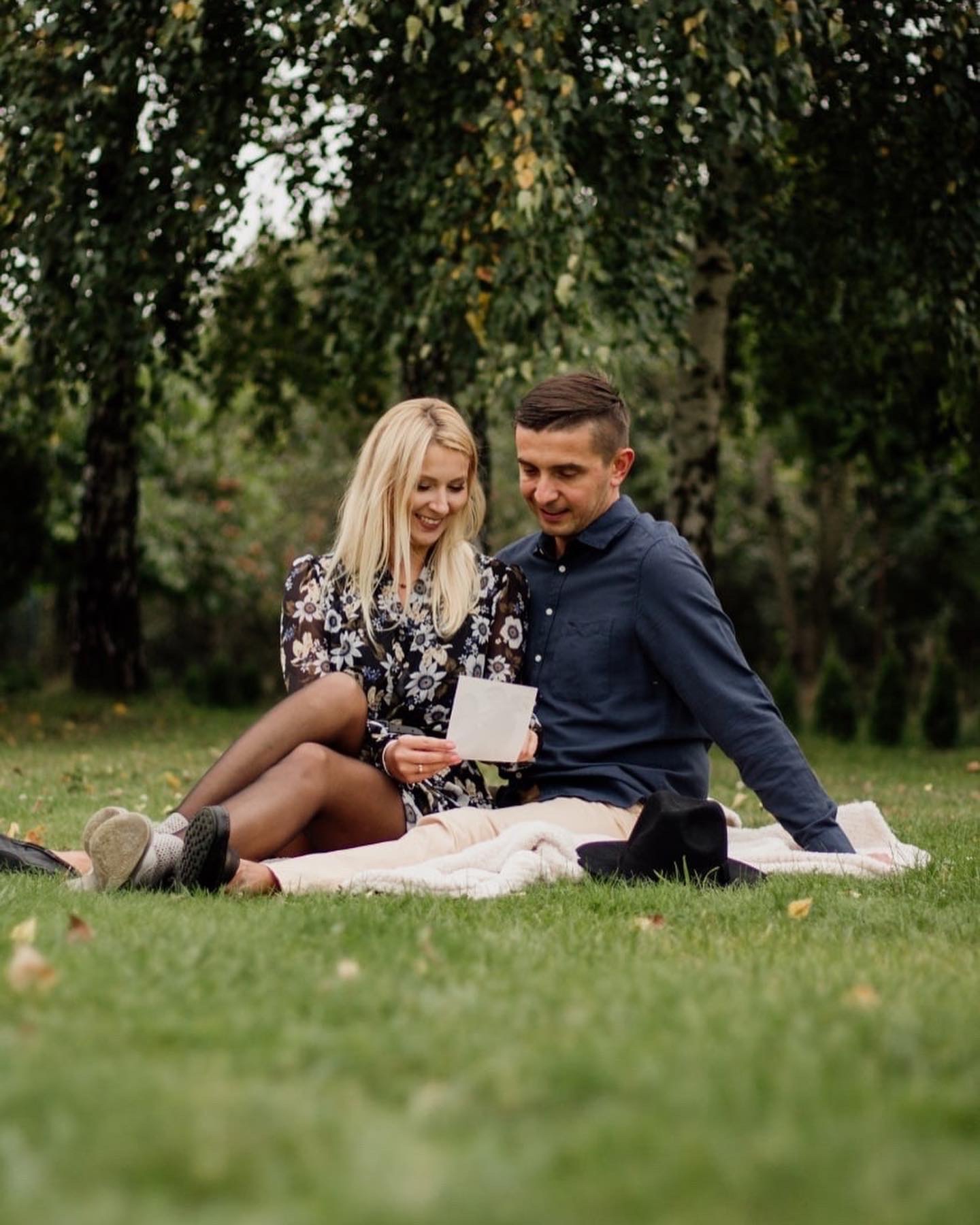 Para siedząca na kocu w parku ogląda zdjęcia, kobieta w kwiecistej sukience i rajstopach, mężczyzna w granatowej koszuli, czarny kapelusz leży obok nich.