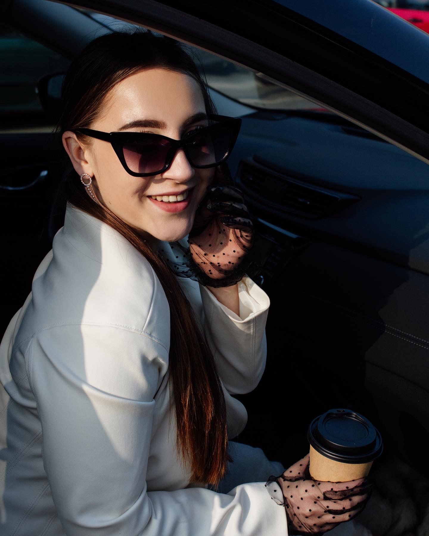 Uśmiechnięta kobieta w okularach przeciwsłonecznych i białej kurtce, siedząca w samochodzie, trzymająca kawę na wynos i rozmawiająca przez telefon