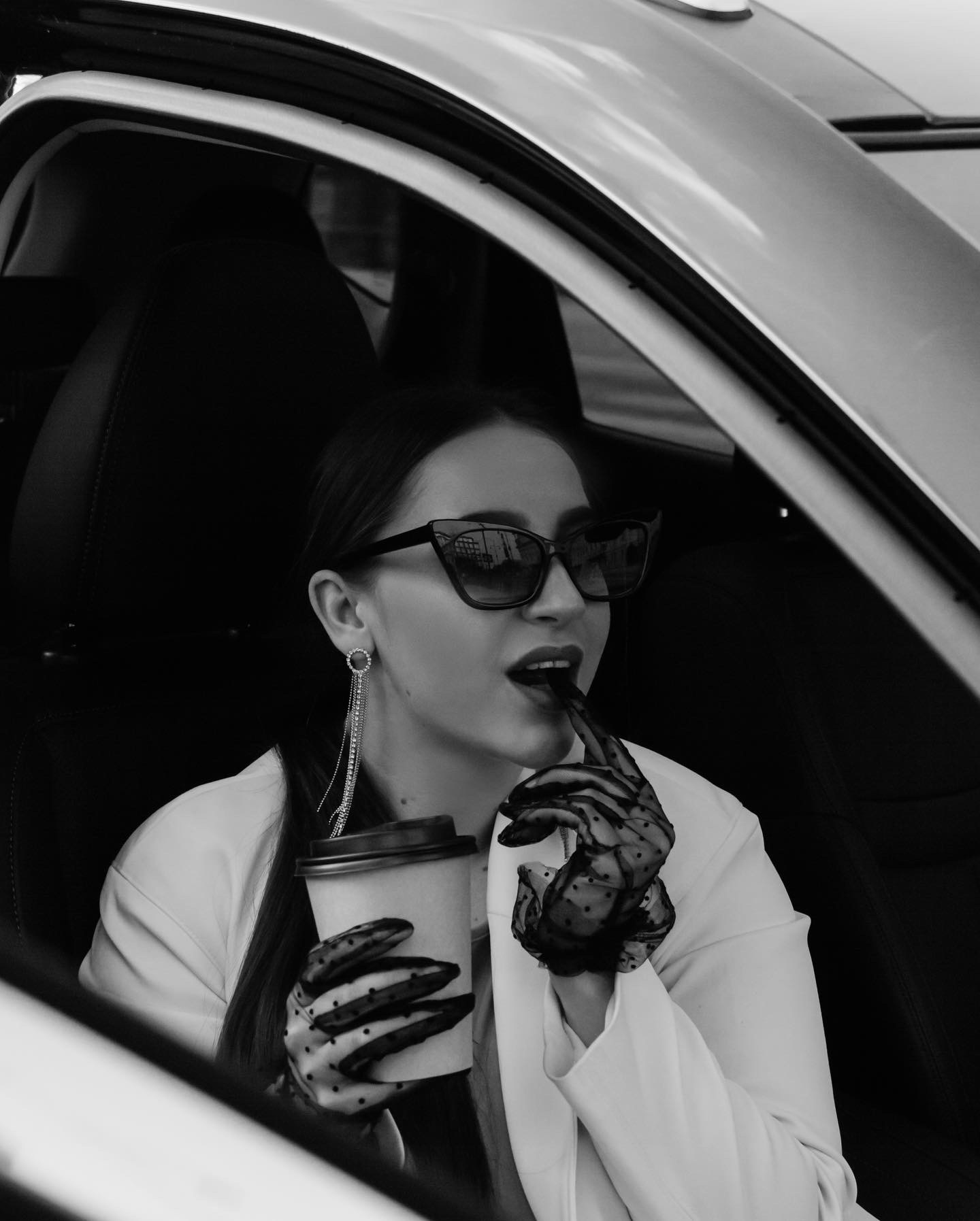 Czarno-białe zdjęcie kobiety w samochodzie, w okularach przeciwsłonecznych i rękawiczkach w kropki, pijącej kawę i trzymającej w ustach czarny długopis.