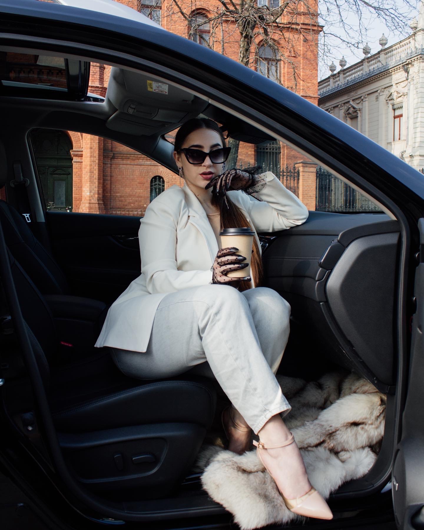 Elegancka kobieta w białym garniturze i okularach przeciwsłonecznych siedzi w czarnym samochodzie z otwartym szyberdachem, trzymając kubek z kawą na tle budynku z czerwonej cegły.