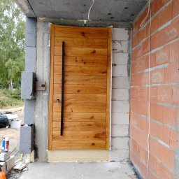 Polska Technika Montażu Drzwi Daniel Kostecki - Nowe drewniane drzwi zewnętrzne z długim, czarnym uchwytem, zamontowane w surowym stanie budynku, z widocznymi bloczkami betonowymi i cegłami.