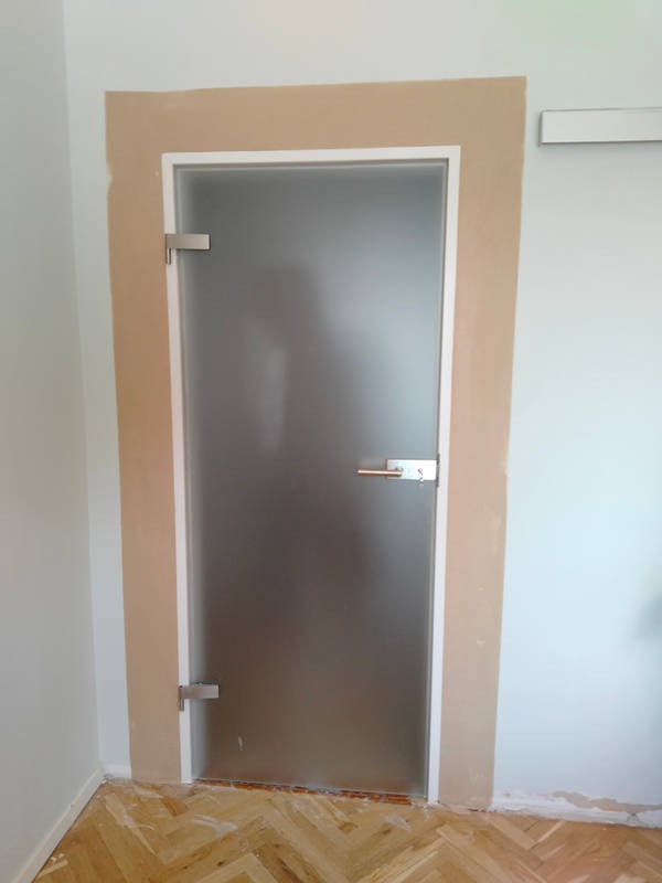 Szklane, matowe drzwi z metalową klamką i zawiasami, zamontowane w ościeżnicy przygotowanej do wykończenia, widoczne na tle parkietu w jodełkę w Chorzowie.