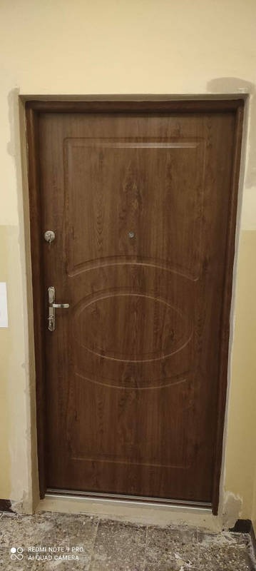 Brązowe drzwi wejściowe z wizjerem i zamkiem, widoczne ślady montażu na ścianie i podłodze.