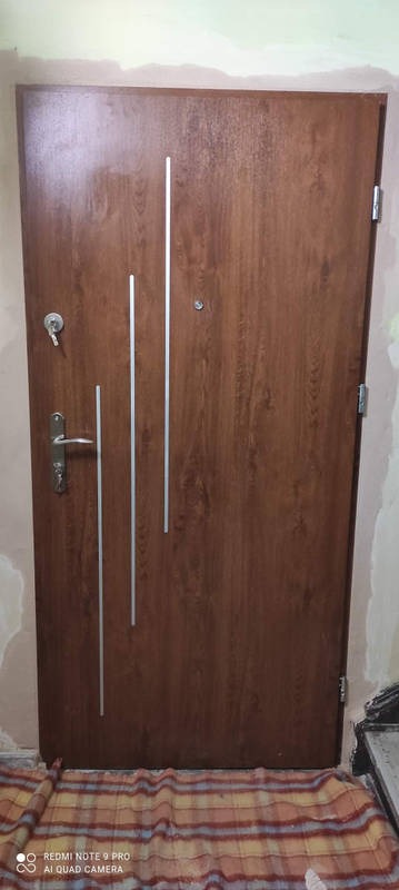 Brązowe drzwi wejściowe z trzema srebrnymi, pionowymi paskami, zamontowane w ościeżnicy w trakcie prac wykończeniowych, widoczny klucz w zamku i fragment niepomalowanej ściany.