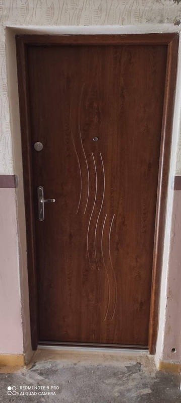 Brązowe drzwi wejściowe z dekoracyjnymi, pionowymi żłobieniami i srebrną klamką, zamontowane w ościeżnicy w budynku mieszkalnym.