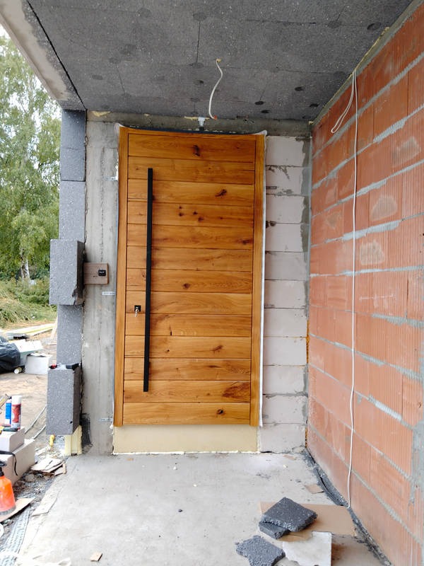 Nowe drewniane drzwi zewnętrzne z długim, czarnym uchwytem, zamontowane w surowym stanie budynku, z widocznymi bloczkami betonowymi i cegłami.