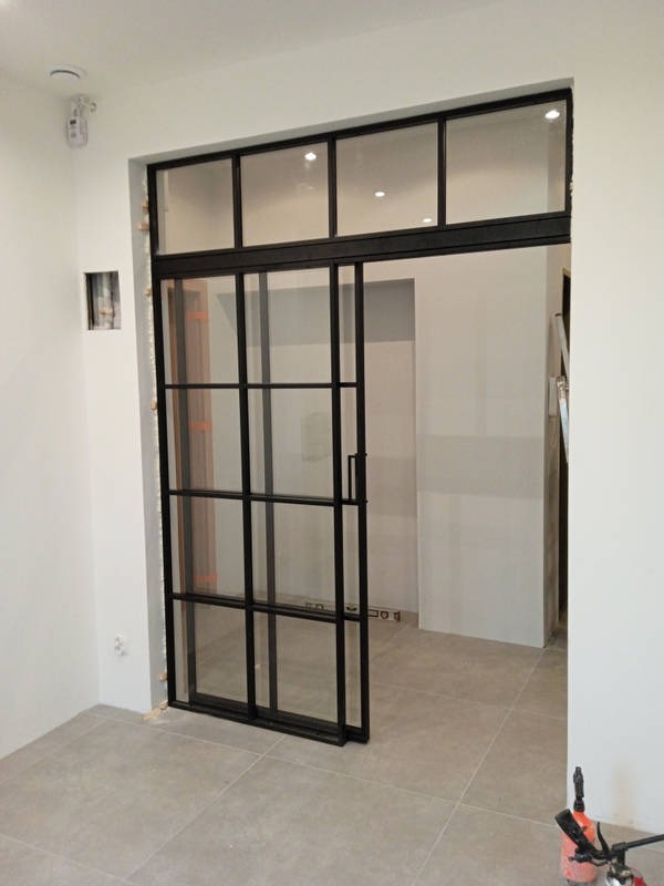 Przesuwne drzwi szklane w stylu loft z czarną ramą, widoczne ślady montażu na ścianie, szpachla i niedomalowane fragmenty, widok na sąsiednie pomieszczenie z szarą podłogą.