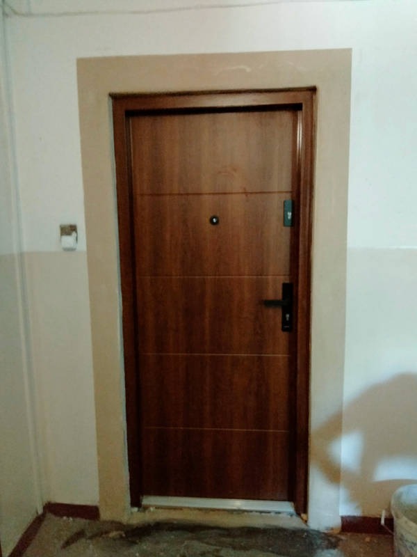 Nowo osadzona, brązowa, metalowa, antywłamaniowa brama wejściowa z czarną klamką i wizjerem, osadzona w ościeżnicy, z widocznymi śladami prac montażowych na ścianie i podłodze.