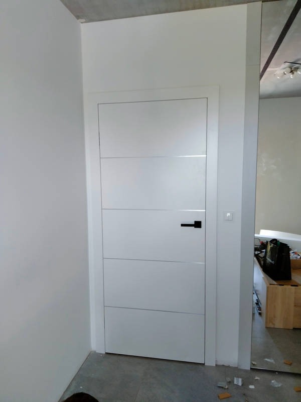 Zamknięte, białe drzwi wewnętrzne z poziomymi frezami i czarną klamką, widoczne ślady montażu na podłodze.