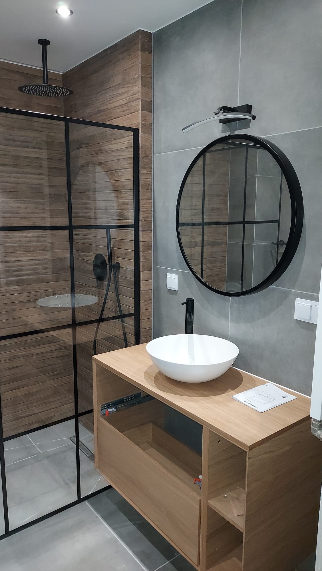 Nowoczesna łazienka z kabiną prysznicową typu walk-in z czarnymi profilami, okrągłym lustrem w czarnej ramie nad umywalką nablatową i drewnianą szafką z otwartą szufladą.