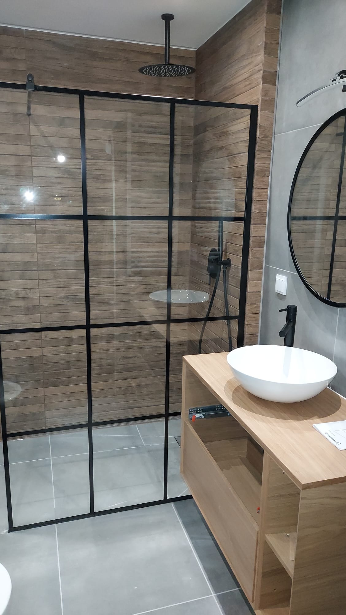 Nowoczesna łazienka z kabiną prysznicową typu walk-in z czarnymi profilami, okrągłym lustrem i umywalką nablatową na drewnianej szafce.