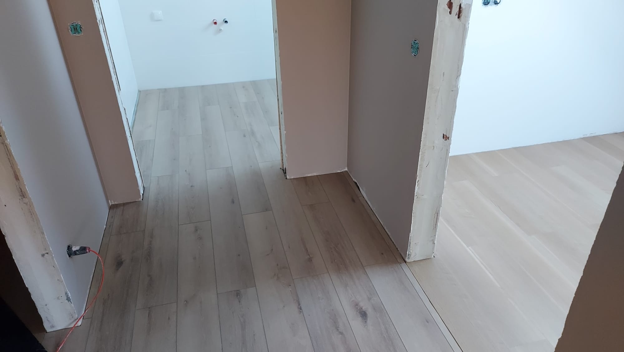 Wnętrze w trakcie wykańczania: jasne panele podłogowe, pomalowane ściany w odcieniach bieli i beżu, widoczne puszki elektryczne i przewody, surowe ościeżnice drzwiowe.