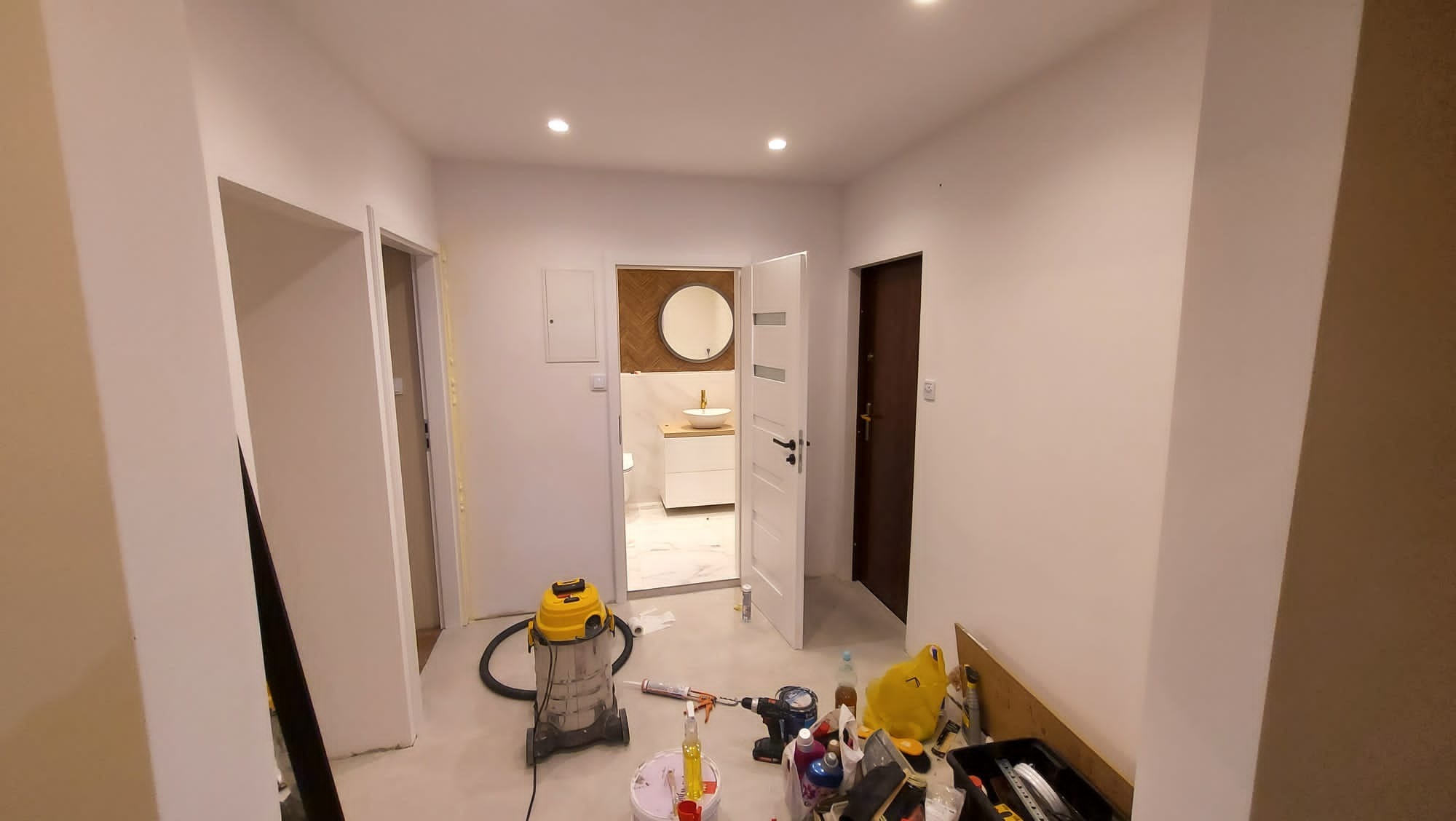 Wnętrze mieszkania w trakcie remontu: widok na odnowioną łazienkę z okrągłym lustrem, białą umywalką i płytkami imitującymi drewno, białe ściany w korytarzu, narzędzia i odkurzacz budowlany...
