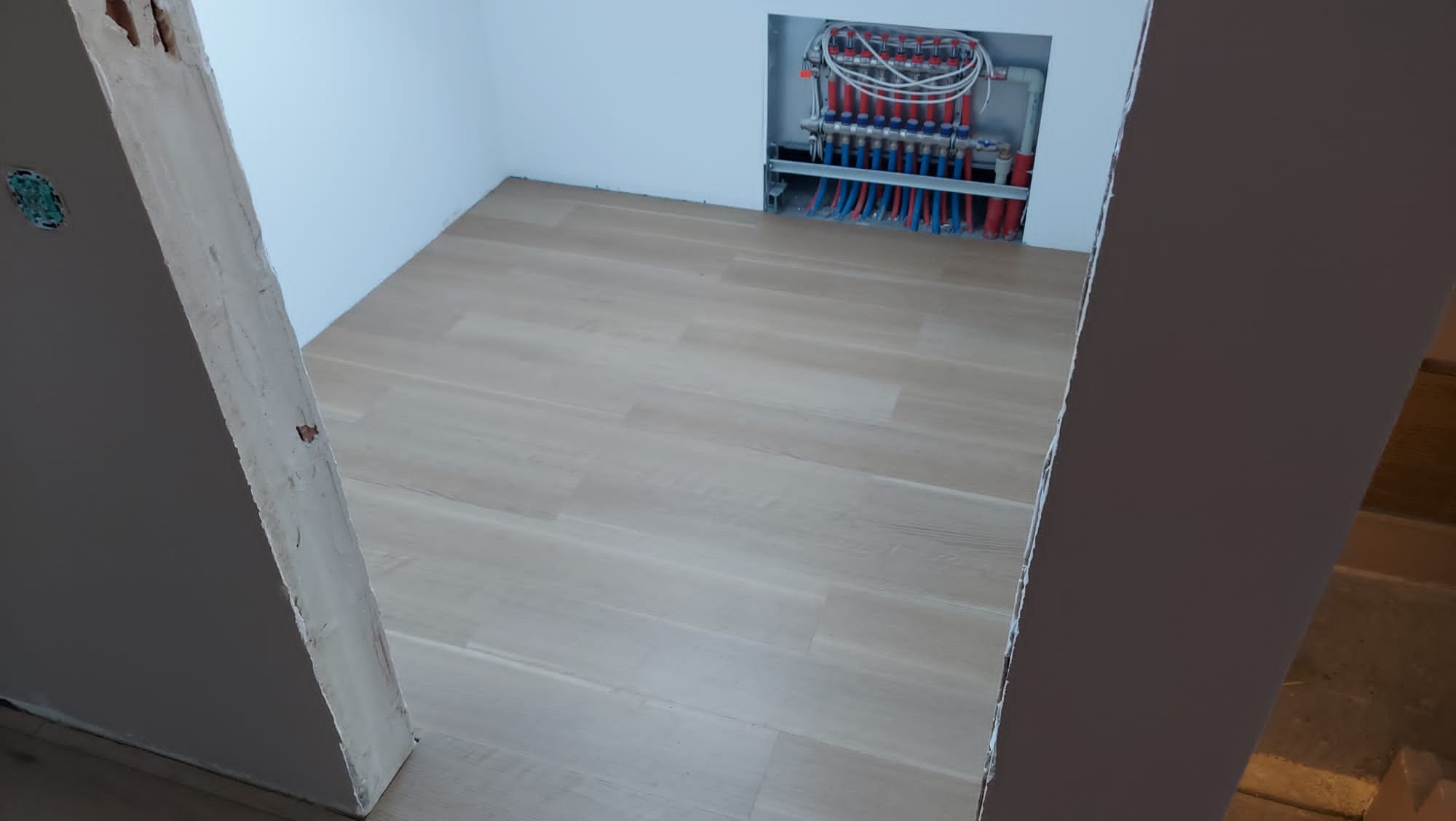 Jasne panele podłogowe w nowym pomieszczeniu, widoczna instalacja ogrzewania podłogowego w ścianie, surowe wykończenie ościeżnicy.