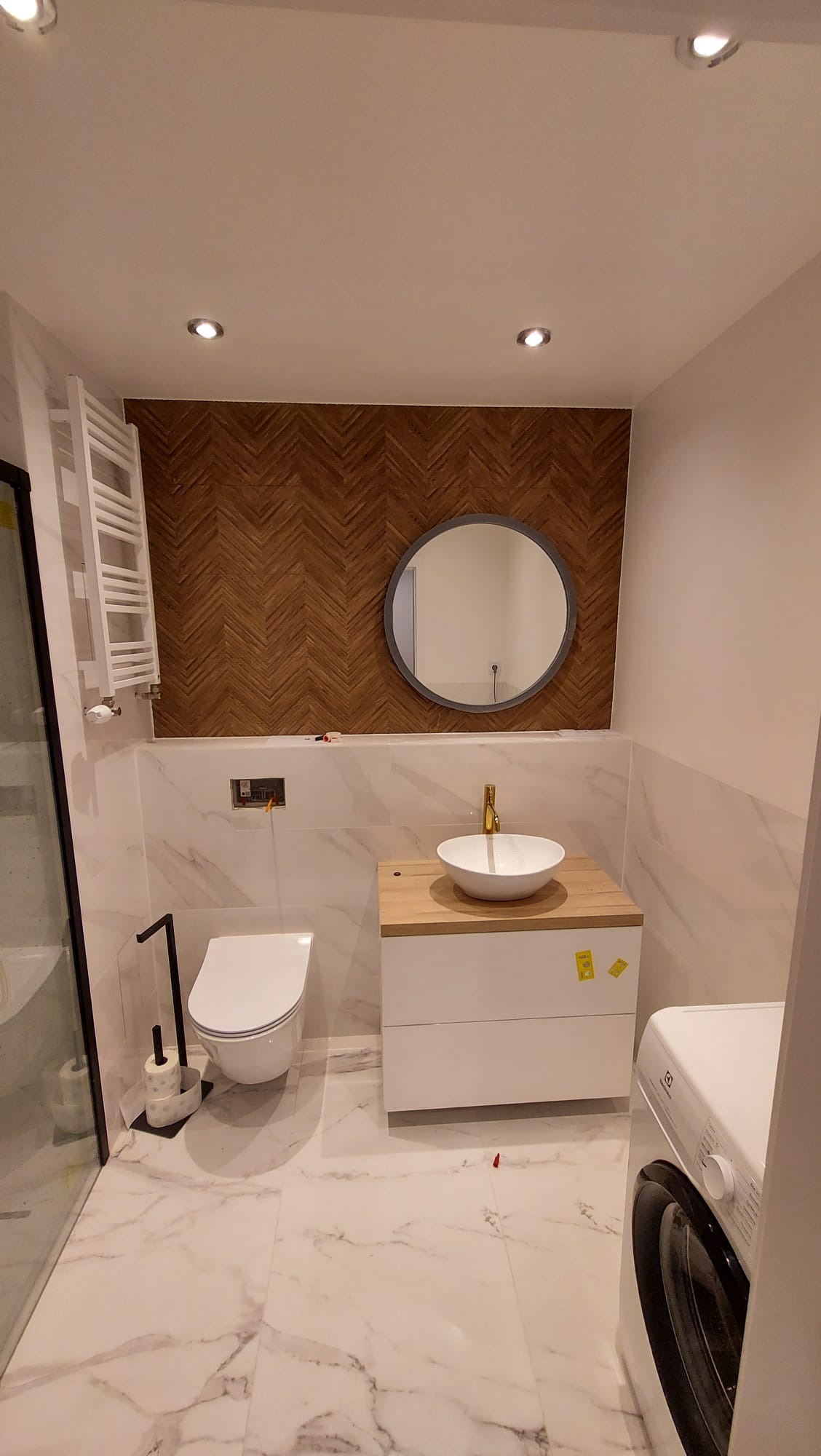 Nowoczesna łazienka z drewnianą ścianą w jodełkę, okrągłym lustrem, białą umywalką na drewnianym blacie, marmurową podłogą i pralką Electrolux.