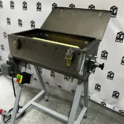 Metalowa skrzynia z otwieraną pokrywą, zamontowana na stalowym stelażu z silnikiem elektrycznym i panelem sterowania, na tle ściany z powtarzalnym logo spawalniczym.