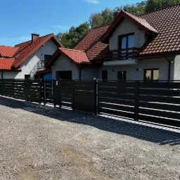 Nowoczesna, pozioma, czarna brama wjazdowa z furtką, widoczna na tle dwóch domów z czerwoną dachówką i szarą elewacją, otoczonych zielenią.