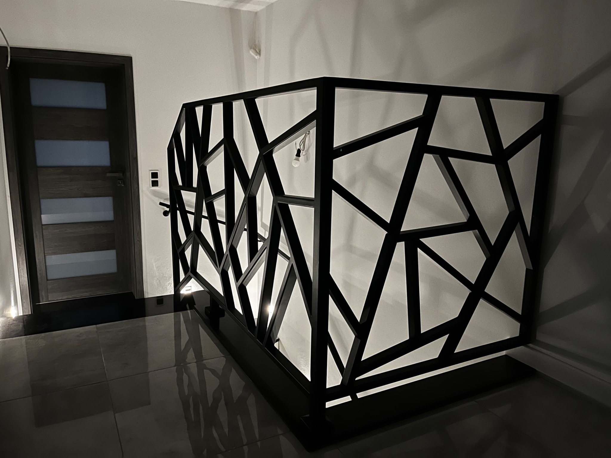 Nowoczesna balustrada schodowa z czarnego metalu o geometrycznym wzorze, widoczna na tle białej ściany i ciemnej podłogi, z dekoracyjną żarówką zwisającą z górnej krawędzi.