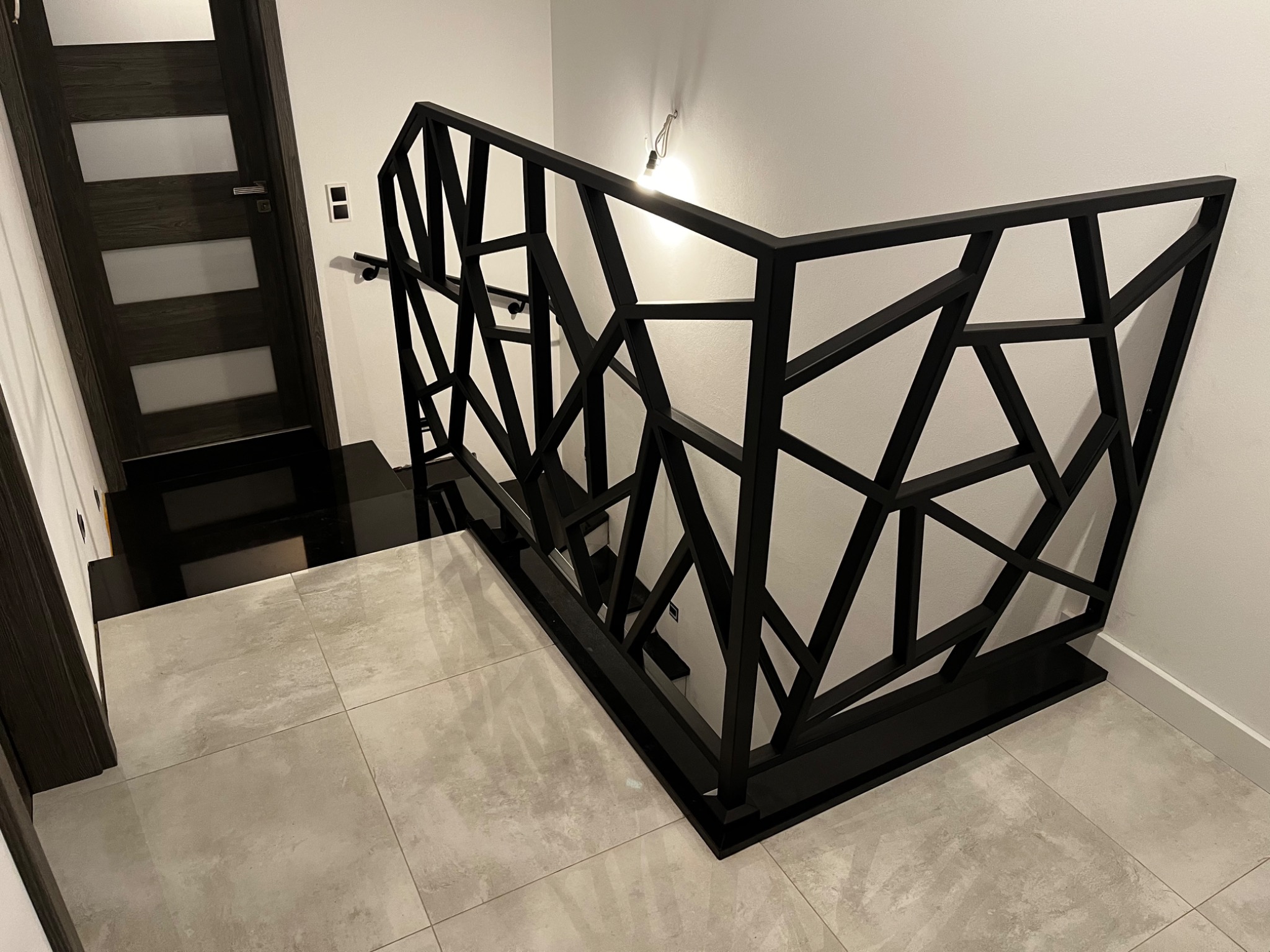 Nowoczesna, czarna balustrada schodowa o geometrycznym wzorze, wykonana z metalu, na tle jasnych płytek podłogowych i ciemnych schodów z drewnianymi drzwiami w tle.