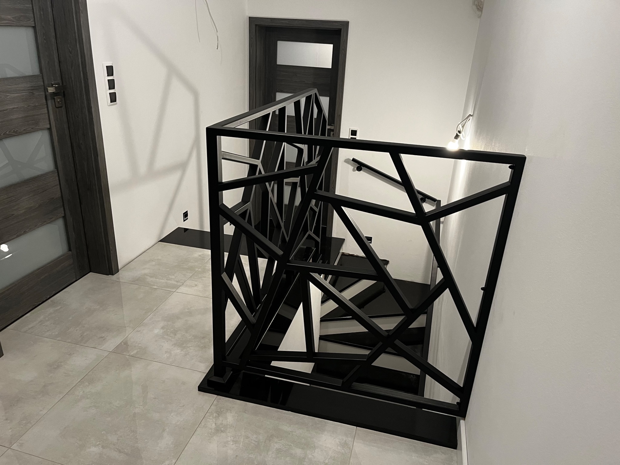 Nowoczesna, czarna balustrada schodowa o geometrycznym wzorze, wykonana z metalu, widoczna z góry na tle białych ścian i szarych płytek podłogowych, z widocznymi schodami w czarno-białej kolorystyce.