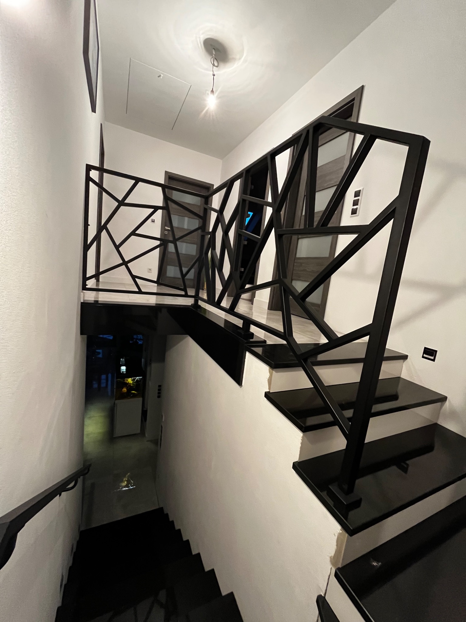 Nowoczesne schody z czarnymi stopniami i oryginalną, geometryczną balustradą w ciemnym kolorze, widok z góry na dół z elementami wystroju wnętrza w tle.