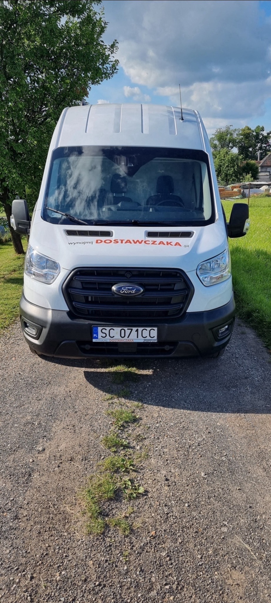 Biały Ford Transit z napisem 'wynajmij DOSTAWCZAKA.pl' na przedniej szybie, zaparkowany na żwirowej drodze z kępami trawy, widok z przodu.
