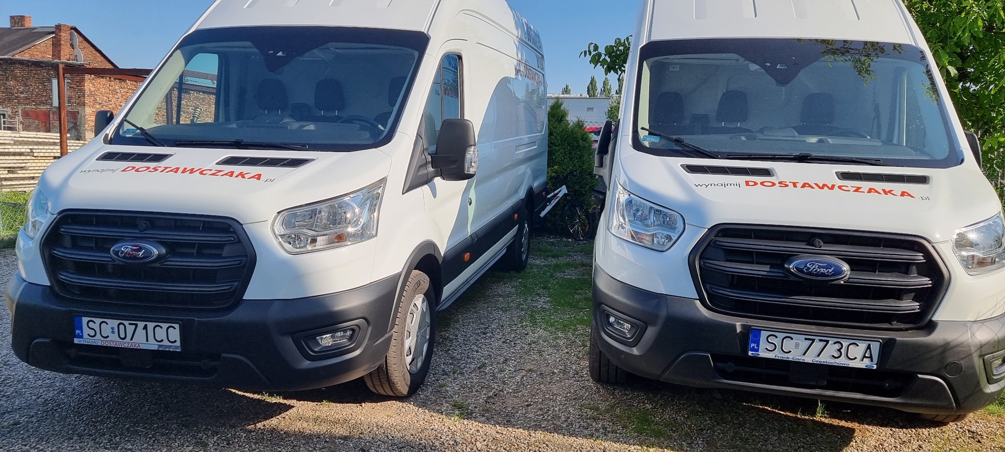 Dwa białe busy Ford Transit z napisem 'wynajmij DOSTAWCZAKA.pl' zaparkowane obok siebie na żwirowym podłożu, z widocznymi numerami rejestracyjnymi SC-071CC i SC-773CA.