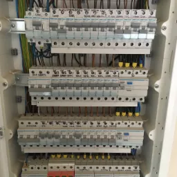 Rozdzielnica elektryczna z zainstalowanymi bezpiecznikami i okablowaniem, widoczne opisy obwodów pisane markerem na obudowach aparatów, jeden z wyłączników ze świecącą diodą sygnalizującą stan.