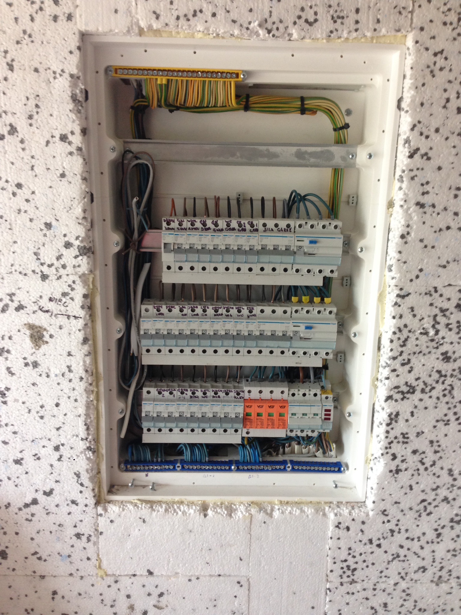 Rozdzielnica elektryczna z bezpiecznikami i okablowaniem, zamontowana w ścianie z ociepleniem styropianowym, widoczne napisy markerem na urządzeniach i ścianie.