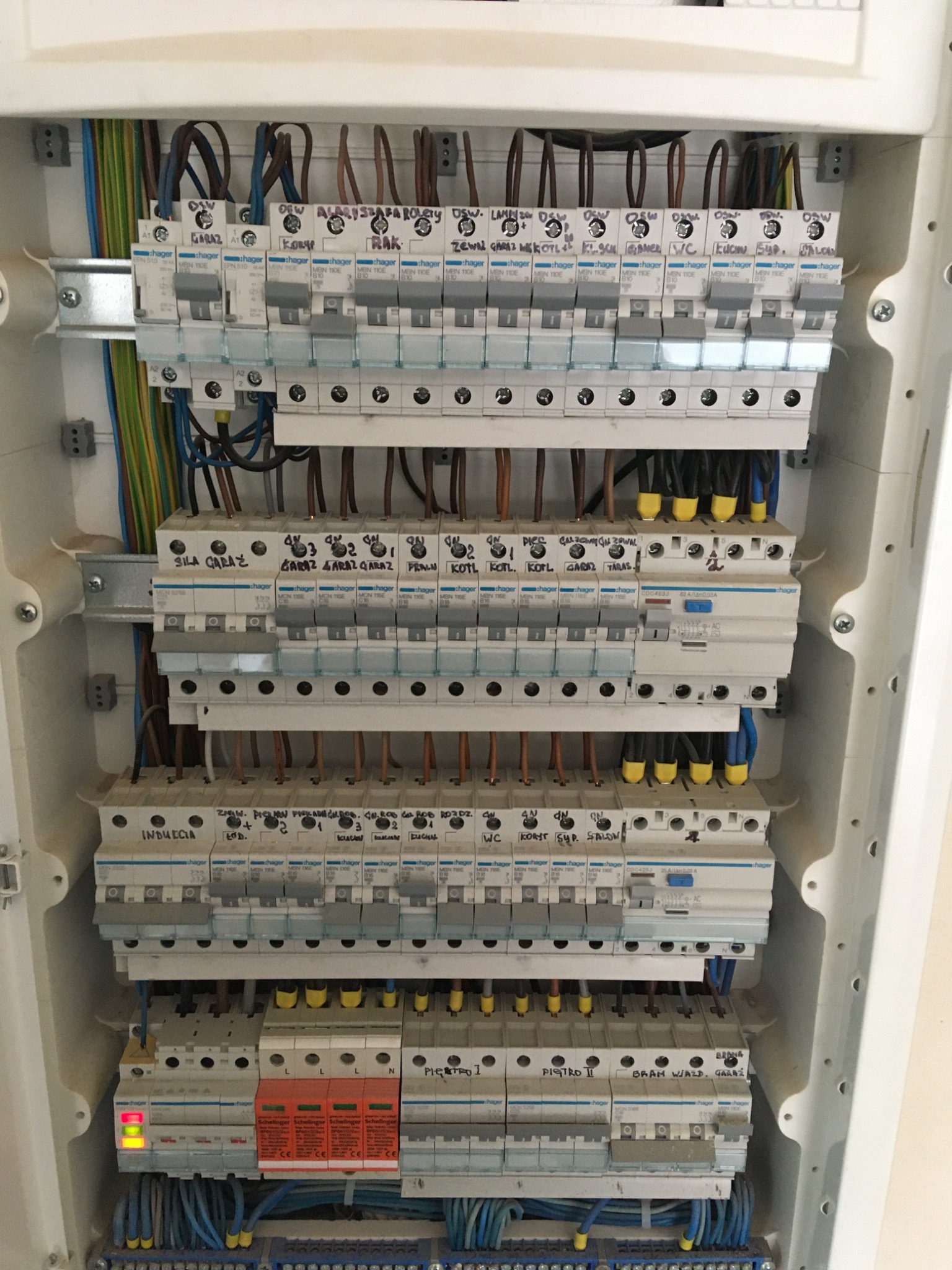 Rozdzielnica elektryczna z zainstalowanymi bezpiecznikami i okablowaniem, widoczne opisy obwodów pisane markerem na obudowach aparatów, jeden z wyłączników ze świecącą diodą sygnalizującą stan.