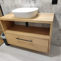 Drewniana szafka łazienkowa z umywalką nablatową, widoczna szuflada z czarnym uchwytem, wzorzyste płytki na ścianie w tle.