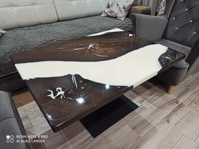 Stolik kawowy z blatem z drewna i żywicy epoksydowej, z wyraźnym podziałem na ciemną część z naturalnym rysunkiem drewna i białą, połyskującą żywicę, ustawiony w salonie obok szarego fotela i sofy.