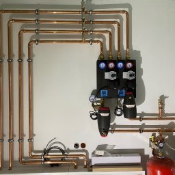 KIS-BUD Rafał Kisilewicz - Rozdzielacz ogrzewania podłogowego z miedzianymi rurami i pompami obiegowymi Flamco zamontowany na białej ścianie, widoczne manometry i zawory.