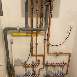 KIS-BUD Rafał Kisilewicz - Rozdzielacz ogrzewania podłogowego z piecem gazowym, widoczne miedziane rury i szare rury PCV, żółty przewód gazowy, zamontowane na białej ścianie.
