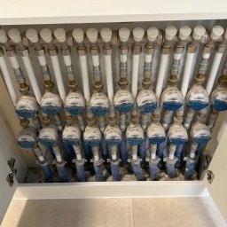 KIS-BUD Rafał Kisilewicz - Rozdzielacz ogrzewania podłogowego w szafce, widoczne liczniki z niebieskimi oznaczeniami i numerami pętli grzewczych, białe rury i izolacja.