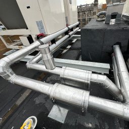 KIS-BUD Rafał Kisilewicz - Metalowe rury wentylacyjne na dachu budynku, widoczne elementy systemu wentylacji mechanicznej z odzyskiem ciepła oraz fragment dachu z papy i dachówki.