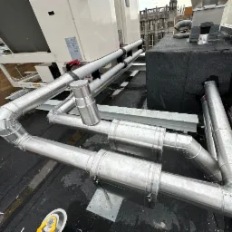 Metalowe rury wentylacyjne na dachu budynku, widoczne elementy systemu wentylacji mechanicznej z odzyskiem ciepła oraz fragment dachu z papy i dachówki.