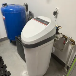 Biały zmiękczacz wody Viessmann z szarym paskiem, podłączony do instalacji hydraulicznej, obok niebieski zbiornik ciśnieniowy, widoczne zasilanie z gniazdka elektrycznego.
